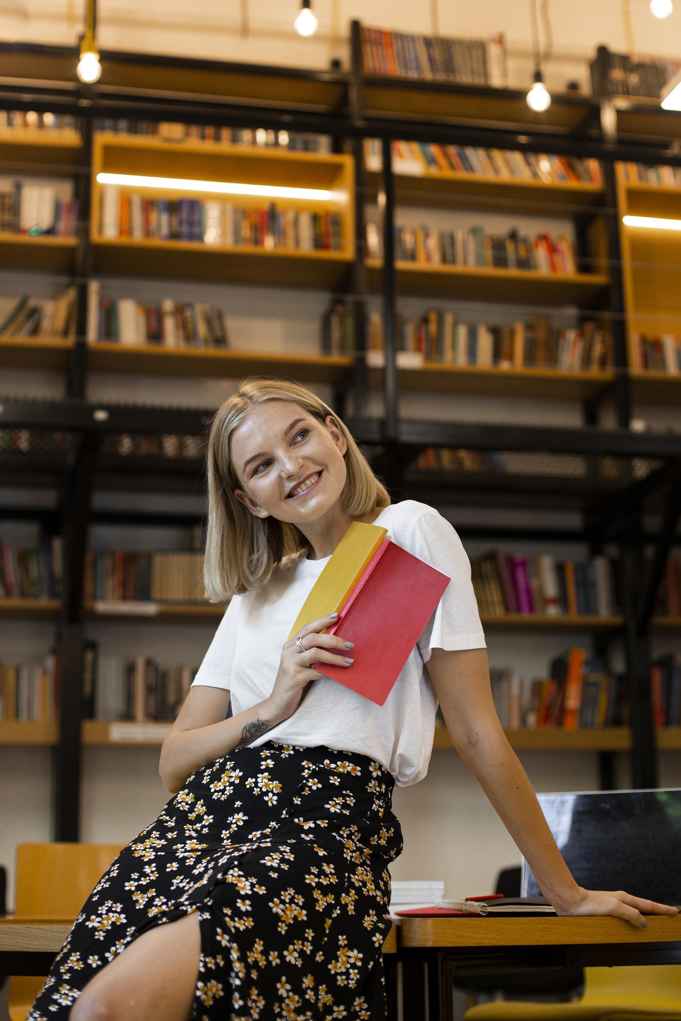 Uma mulher sorridente sentada em uma mesa de biblioteca, segurando dois livros de capa colorida, estantes cheias ao fundo