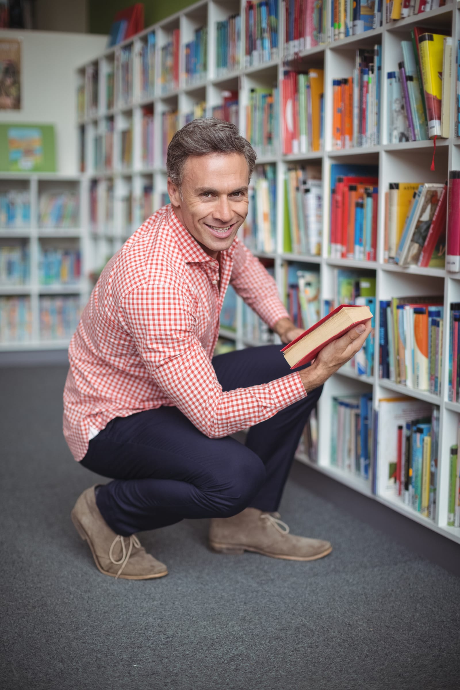 Um homem agachado sorrindo, segurando um livro de capa vermelha diante de estantes cheias de livros