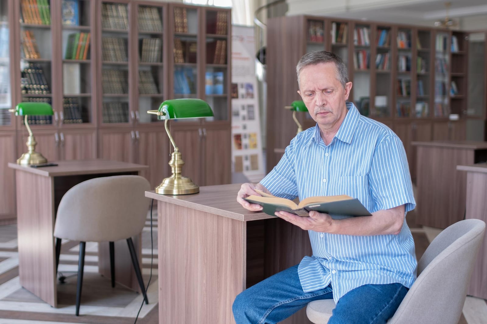Um homem sentado à mesa de uma biblioteca, lendo um livro encadernado sob luz de luminária verde