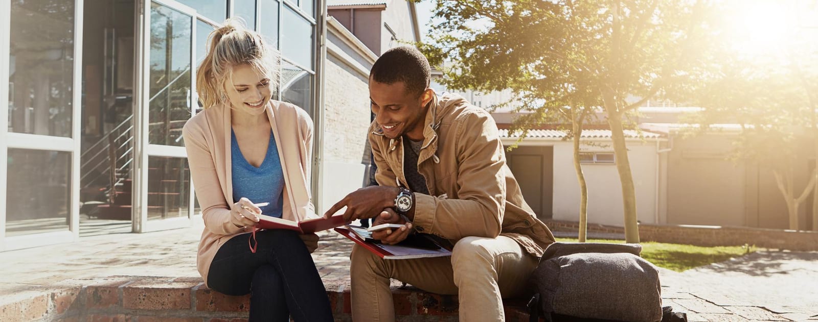 Dois jovens sentados ao ar livre, lendo um livro de capa vermelha e conversando